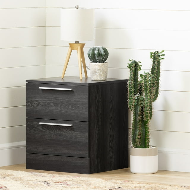 South Shore Step One Essential 2-Drawer Nightstand for Adult in gray oak finish with two large drawers and slim silver handles, shown front-facing on a light wood floor against white shiplap walls, topped with a wooden tripod lamp with a white shade and a small round cactus in a white textured pot, beside a tall green cactus in a white and beige planter, ideal for minimalist bedroom storage and organization.