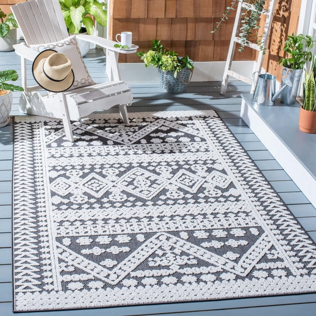 SAFAVIEH Cabana Arielle Geometric Indoor/Outdoor Area Rug with black and white diamond and floral patterns on a grey wooden deck, next to a white Adirondack chair with a beige pillow and straw hat, surrounded by potted plants and metal watering cans, suitable for busy indoor or outdoor areas.