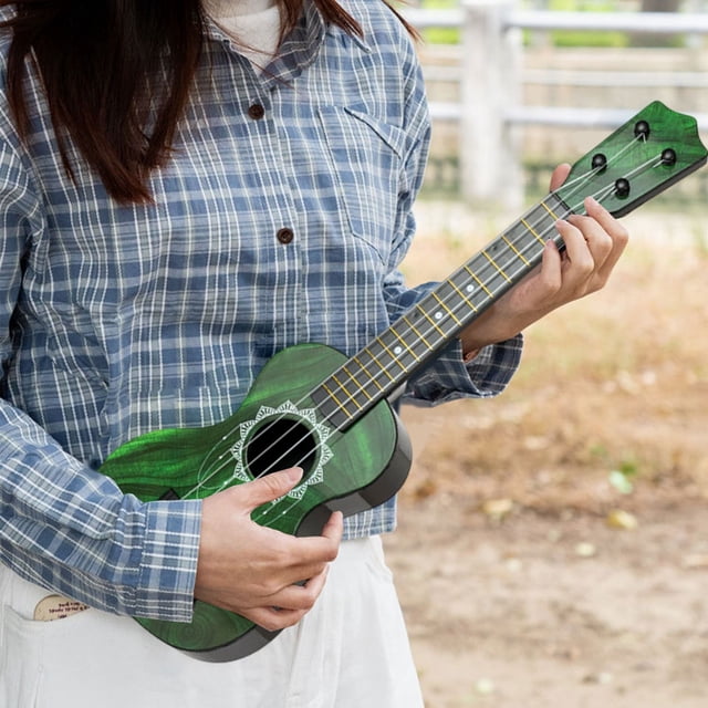 Clearance! JMYHOAM Children's Hippopotamus Pattern Guitar Ukulele Simulation Playable Instrument Early Education Enlightenment Toy. A green ukulele with a hippopotamus pattern, designed for children, held by a person outdoors. The instrument features a medium size suitable for small hands, durable plastic material, and comes with a random color pick. Ideal for early music education and hand-eye coordination development.