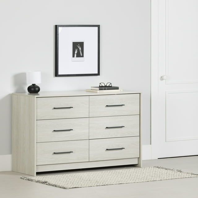 South Shore Londen 6-Drawer Dresser for Adult in winter oak finish with six spacious drawers featuring long black metal handles and metal slides, shown in a minimalist bedroom setting with a black lamp, stacked books, and framed black-and-white hand photo above, placed on a light woven rug, ideal for organized clothing storage.