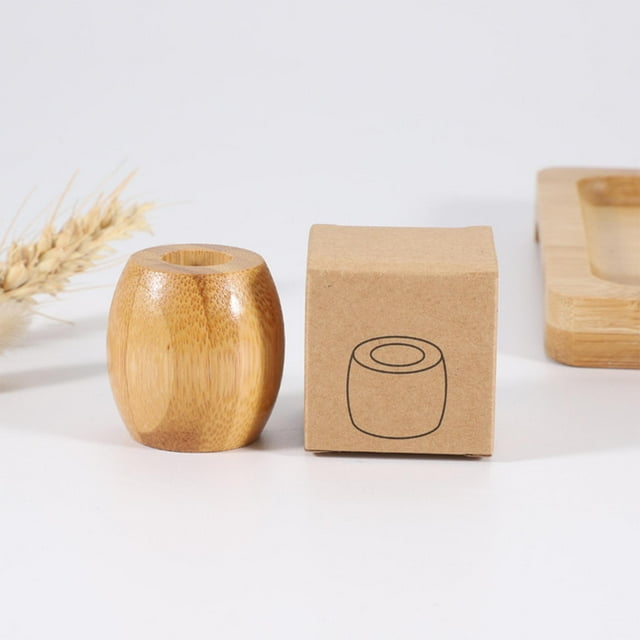 soporte de bambú macarena para cepillos de dientes portalápices encimera, vista frontal centrada de un soporte pequeño de bambú natural de forma ligeramente ovalada con agujero superior circular y vetas de madera visibles; caja de cartón junto a la pieza y bandeja desenfocada al fondo; altura aprox. 4 cm; a menudo usado para sostener cepillos de dientes, bolígrafos o brochas de maquillaje.