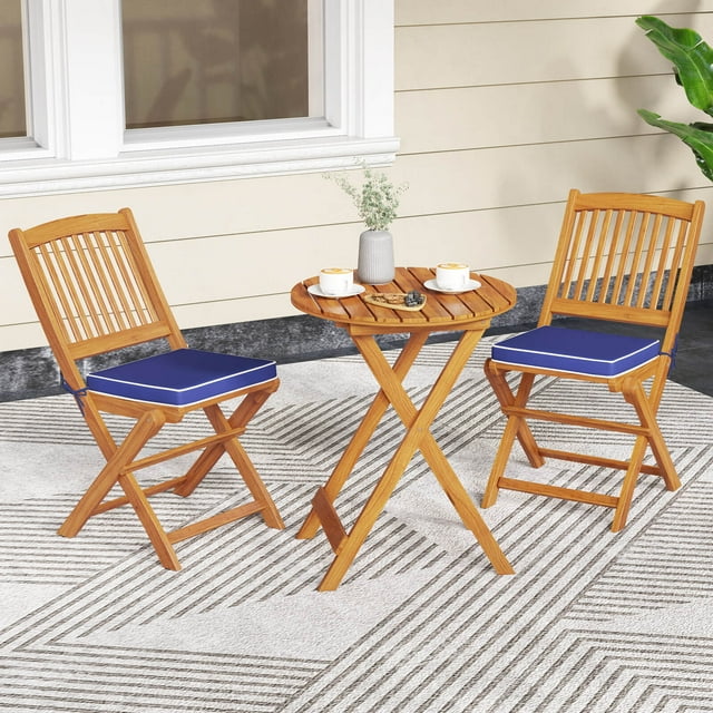 Costway 3 PCS Acacia Wood Patio Bistro Set with a folding round table and two chairs featuring navy cushions. Crafted from solid acacia wood, this compact bistro patio set is perfect for small outdoor spaces like balconies or gardens. The ergonomic chairs have slatted backs and removable cushions for comfort, while the natural wood finish adds rustic charm. Ideal for outdoor dining or relaxing.