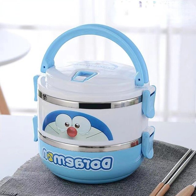 fiambrera térmica de hello kitty para niños — fiambrera portátil de alta capacidad de acero inoxidable con doble compartimento en vista frontal 3/4, acabado blanco y azul con estampado de personaje anime, anillos metálicos pulidos, tapa con asa azul plegable y cierres laterales, apoyada sobre mantel gris junto a palillos; también se usa a menudo para llevar snacks en salidas familiares.