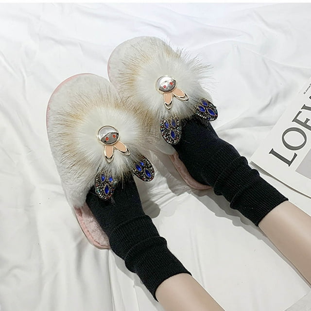 pantuflas de felpa con punta abierta para mujer Rosado, par cenital de sandalias peludas con plantilla y ribete de felpa claro, pompón de pelo sintético blanco con aplique metálico y cristales azules, suela de caucho y tacón bajo 3.2 cm; sobre sábana blanca con pies en calcetas negras.