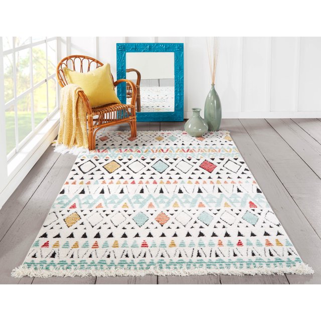 ECARPET Kaia contemporary area rug featuring colorful geometric designs on white background displayed in a bright room with wicker chair, yellow cushion and throw blanket, blue framed mirror leaning against wall, and two green ceramic vases.