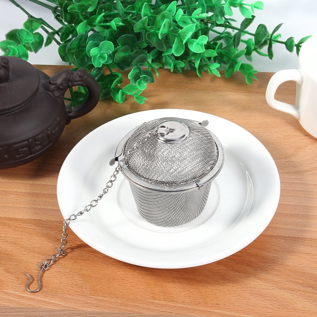 filtro de infusor de malla bola de máquina de té práctica esfera de hierbas colador de especias atrapado en caliente acero inoxidable reticulado ecomeon no Nuevo infusor de té de acero inoxidable 6.5 cm, vista frontal centrada de bola de malla fina con cierre superior y cadena con gancho sobre un platillo blanco; fondo con tetera y planta decorativa, también usado frecuentemente para colar especias en pequeñas preparaciones.