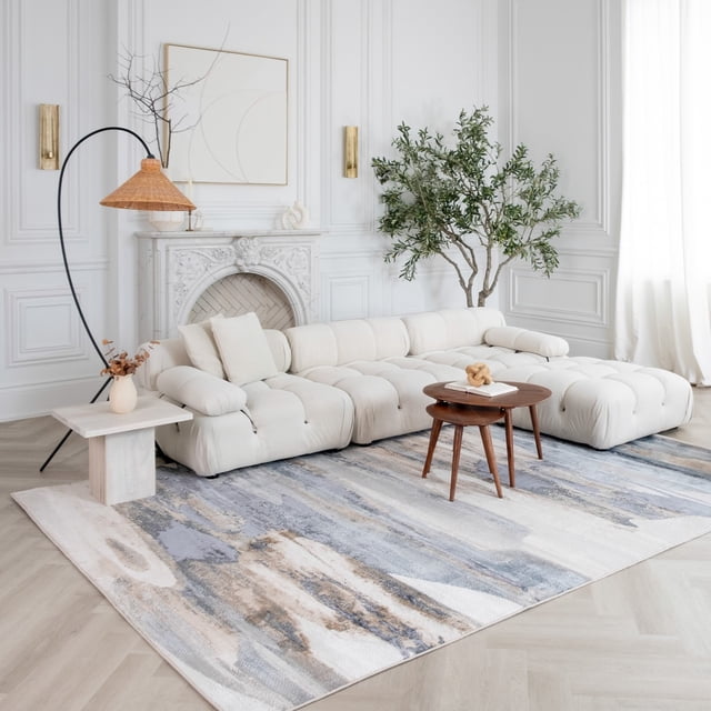 Abstract blue and beige Rug Branch Atlas Mirage indoor area rug placed under a tufted white sectional sofa in an elegant living room featuring minimalist art, wooden coffee table, floor lamp, and potted plant.