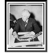 Historic Framed Print, [Frank Lloyd Wright, half-length portrait, seated, facing slightly left, pointing to his design for Crystal Heights].Harris & Ewing., 17-7/8" x 21-7/8"