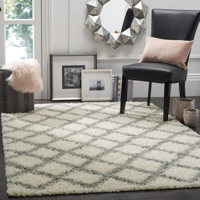Safavieh Dallas Jerrie geometric shag area rug in cream and gray diamond pattern on dark wood floor, styled with black chair, framed photos, decorative mirror, and soft pink pillow.