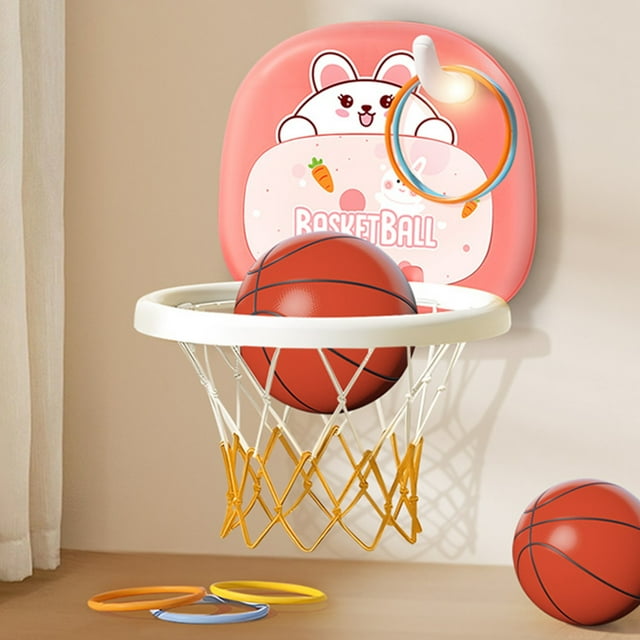 xihbxyly hallazgos de ganga canasta de baloncesto colgante para interiores para bebés 12 a 3 años canasta de baloncesto para uso doméstico juguete de pelota para niños rosa: vista frontal centrada de canasta infantil colgante rosa con respaldo decorado con un conejo, aro blanco con red amarilla y pelota pequeña dentro del aro; otra pelota sobre la mesa y tres aros planos de colores en la superficie; adecuada para juego interior y desarrollo motriz en niños pequeños.