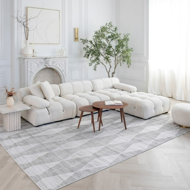 Grey abstract patterned modern indoor area rug placed under a white tufted sectional sofa and wooden coffee tables in a bright, elegant living room.
