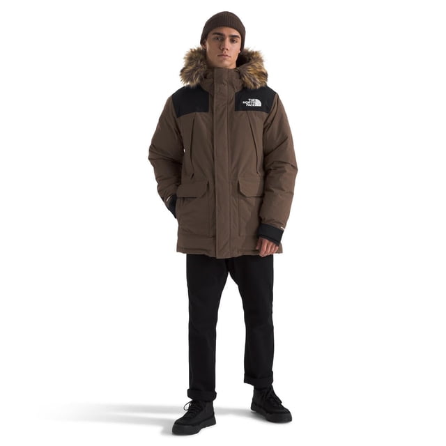 chaqueta parka the north face mcmurdo para hombre smokey brown/tnf black — parka larga frontal marrón ahumado con paneles negros en hombros, capucha con volante de piel sintética extraíble, cierre central con solapa y cremallera, cuatro bolsillos frontales y logo blanco en el pecho; ajuste holgado sobre pantalones negros sobre fondo blanco; adecuada para viajes de invierno por su largo y almacenamiento.
