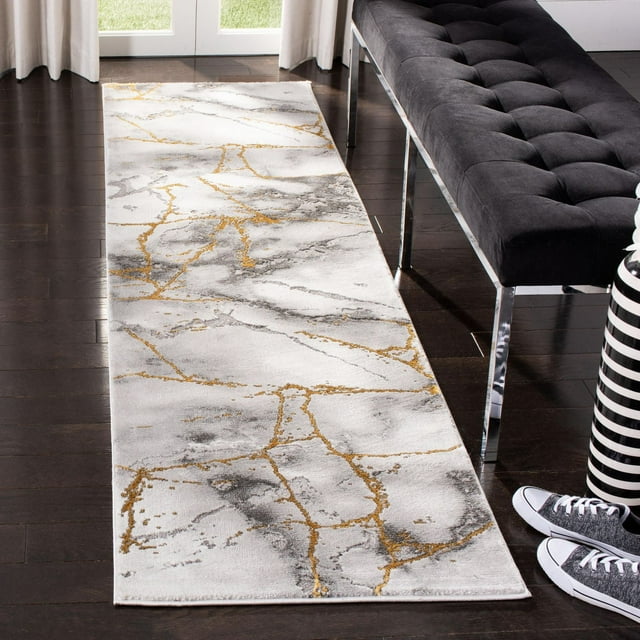 SAFAVIEH Craft Paul abstract marble area rug in white with gold and gray veining displayed on dark wood floor beside a black tufted bench.