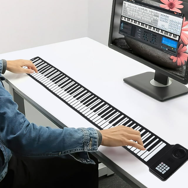 Phenofice Electric Keyboard Silicone Folding Black with Battery, fully extended on a white desk. This portable piano keyboard features soft silicone keys, a built-in speaker, and a compact control panel. Ideal for travel and small spaces, it is shown connected to a computer monitor displaying music software, with a person playing the keys.