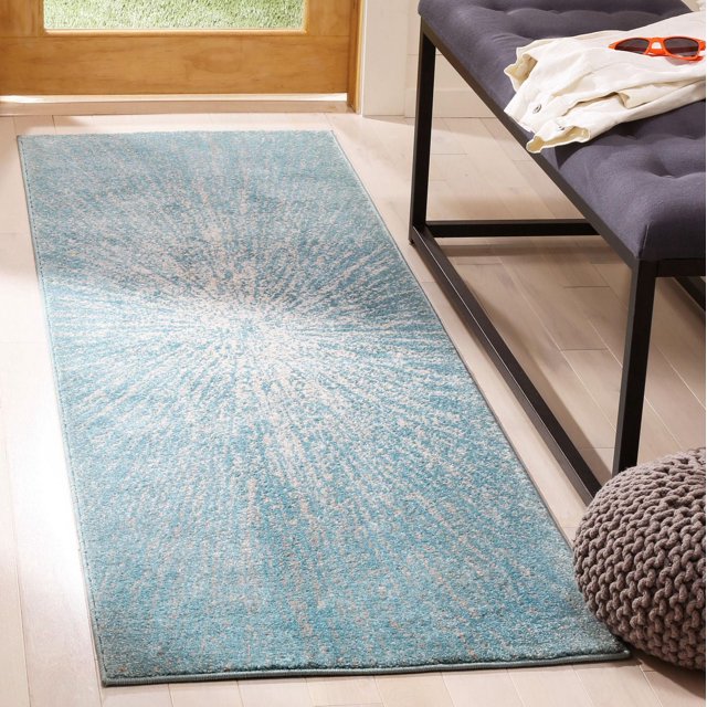 Safavieh Evoke Vana Abstract Geometric Area Rug with a blue and beige radiating sunburst pattern, made of soft cut-pile polypropylene frieze, placed on light hardwood floor near a wooden door and beside a black metal-framed bench with gray tufted cushion and knitted pouf, ideal for entryways and high-traffic areas.
