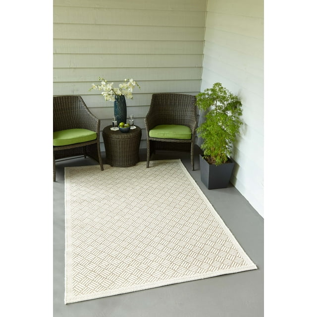 ECARPET Eve Contemporary Area Rug with a tan and ivory geometric basketweave pattern made from stain-resistant polypropylene fibers, shown on a gray porch floor with two brown wicker chairs with green cushions, a matching wicker side table holding a blue vase with white flowers and a bowl of green apples, and a green potted plant in a black planter against white siding walls, ideal for indoor and outdoor spaces.