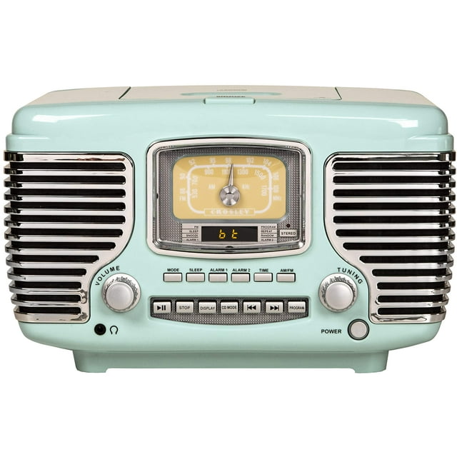 crosley cr612b-ab corsair radio am/fm bluetooth de mesa con reproductor de cd y reloj despertador dual, azul aguamarina vista frontal centrada de la radio Corsair color aqua blue con parrillas laterales cromadas, dial analógico AM/FM amarillo en el centro, pequeña pantalla digital que muestra el modo, perillas rotativas de volumen y sintonía, fila de botones de reproducción y bandeja superior para CD; fondo blanco. Su tamaño compacto la hace adecuada para mesa de noche o repisa y a menudo se usa como radio despertador decorativo.