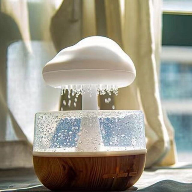 humidificador de lluvia con ruido blanco, humidificador nube en forma de hongo colorida gota de agua para dormir — vista frontal centrada de un humidificador blanco con tapa en forma de nube/hongo sobre base cilíndrica con acabado imitación madera y depósito translúcido con gotas; emite finas gotas de vapor que simulan lluvia y tiene botones visibles en la base. Para uso doméstico y dormitorio; a menudo usado también como luz nocturna decorativa.