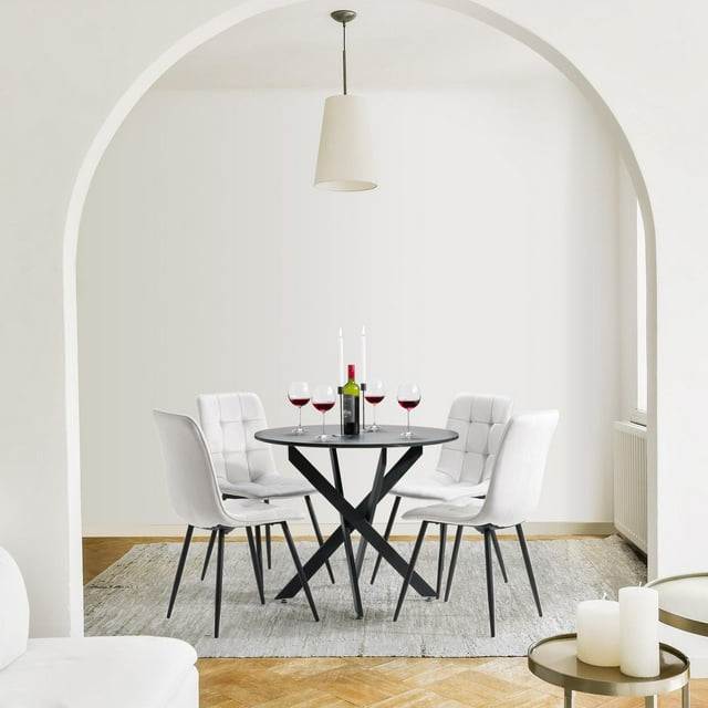 Compact modern round black indoor dining table set for four with white velvet upholstered chairs, styled with wine glasses and candles in a minimalist room.