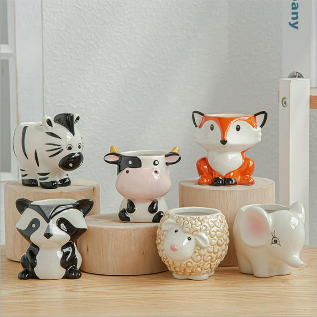 macetas de cerámica con forma de animal, lindas macetas de cerámica con forma de animal, adornos de jardín, máxima comodidad Vista 3/4 de seis macetas esmaltadas: zorro naranja, elefante gris, oveja crema texturizada, vaca blanco y negro, mapache bicolor y cebra rayada, colocadas sobre bases de madera en fondo interior neutro; ideales para suculentas y decoración de patio.