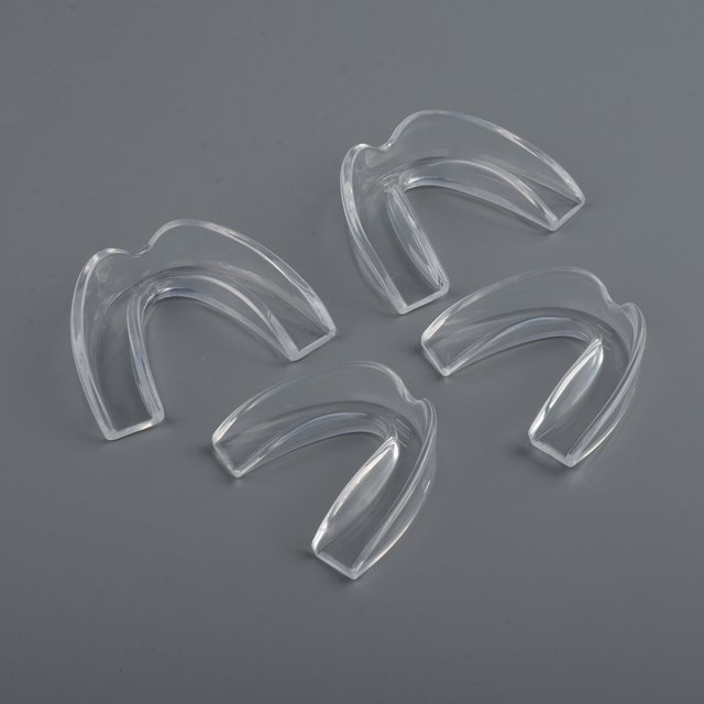 protector bucal para niños para rechinar los dientes para bruxismo, bandeja moldeable transparente 60 x 40 x 25 mm, cuatro protectores dispuestos sobre fondo gris, forma en U con cavidad para la arcada dental y borde biselado. Adecuado para uso nocturno, como bandeja de blanqueamiento y para deportes; a menudo usado para almacenar el protector en viajes.