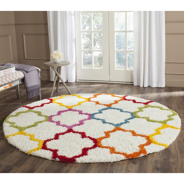 Round Safavieh Shag Kids Yadira area rug featuring a multicolored geometric quatrefoil pattern on white background, placed on wooden floor near window.