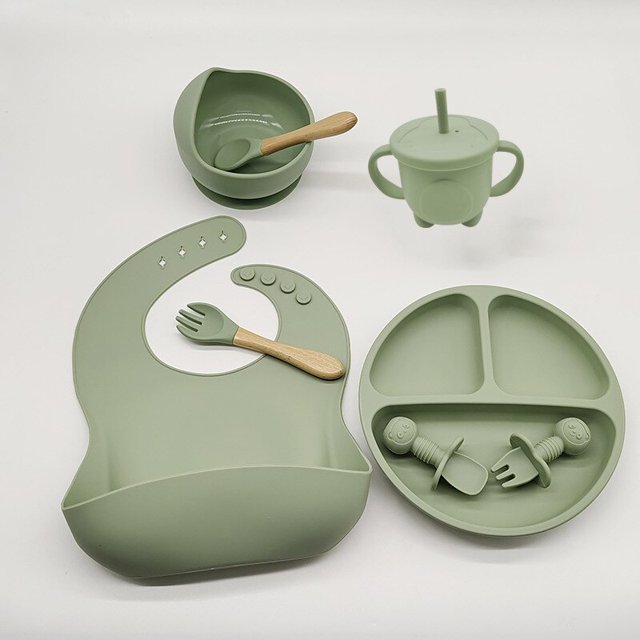 vajilla de silicona de 6-8 piezas para bebé juego de baberos con ventosa tazón cuchara tenedor taza de alimentación plato dividido platos infantiles vajilla para niños suministros para madres y bebés juego de 8 piezas color verde militar: babero 23x30 cm con bolsillo, plato dividido 20x20x3.5 cm con ventosa, cuenco 12x8.5 cm, cuchara y tenedor con mangos de madera y asas cortas, taza con pajilla; vista frontal centrada sobre fondo neutro, acabado liso de silicona de grado alimenticio, ideal para la transición a la autoalimentación y a menudo usado para introducir alimentos sólidos.