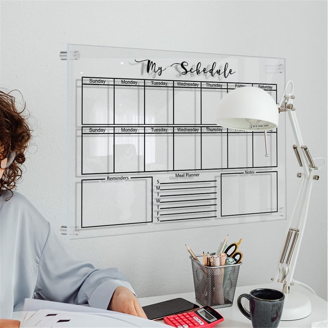 pizarra de acrílico para planificador semanal de pared giyblacko, con borrado en seco transparente, reutilizable, para lista de tareas diarias y semanales. Vista frontal centrada de la pizarra acrílica transparente montada en la pared con cuadrícula semanal visible y secciones para recordatorios, plan de comidas y notas; fijaciones en las esquinas. Escritorio con lámpara, portalápices y taza en primer plano; persona parcialmente visible a la izquierda. También adecuada como organizador de tareas para estudiantes y oficina.