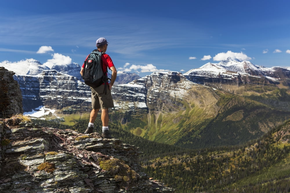 Male hiker standing on top of mountain ridge overlooking snow peaked mountains and forest valley below with blue sky and clouds Waterton Alberta Canada Canvas Art - Michael Interisano Design Pics (19