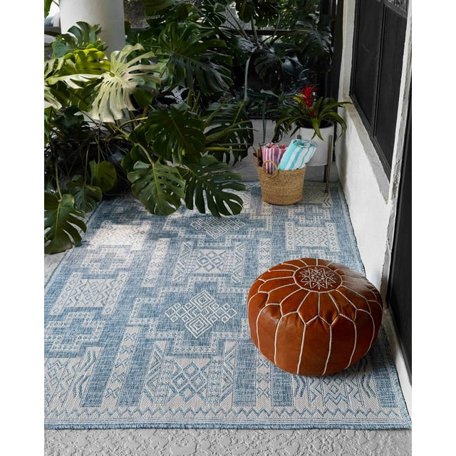 Blue and beige ECARPET Nyra transitional area rug featuring geometric designs in an indoor setting with green plants, brown leather pouf, and basket of towels.