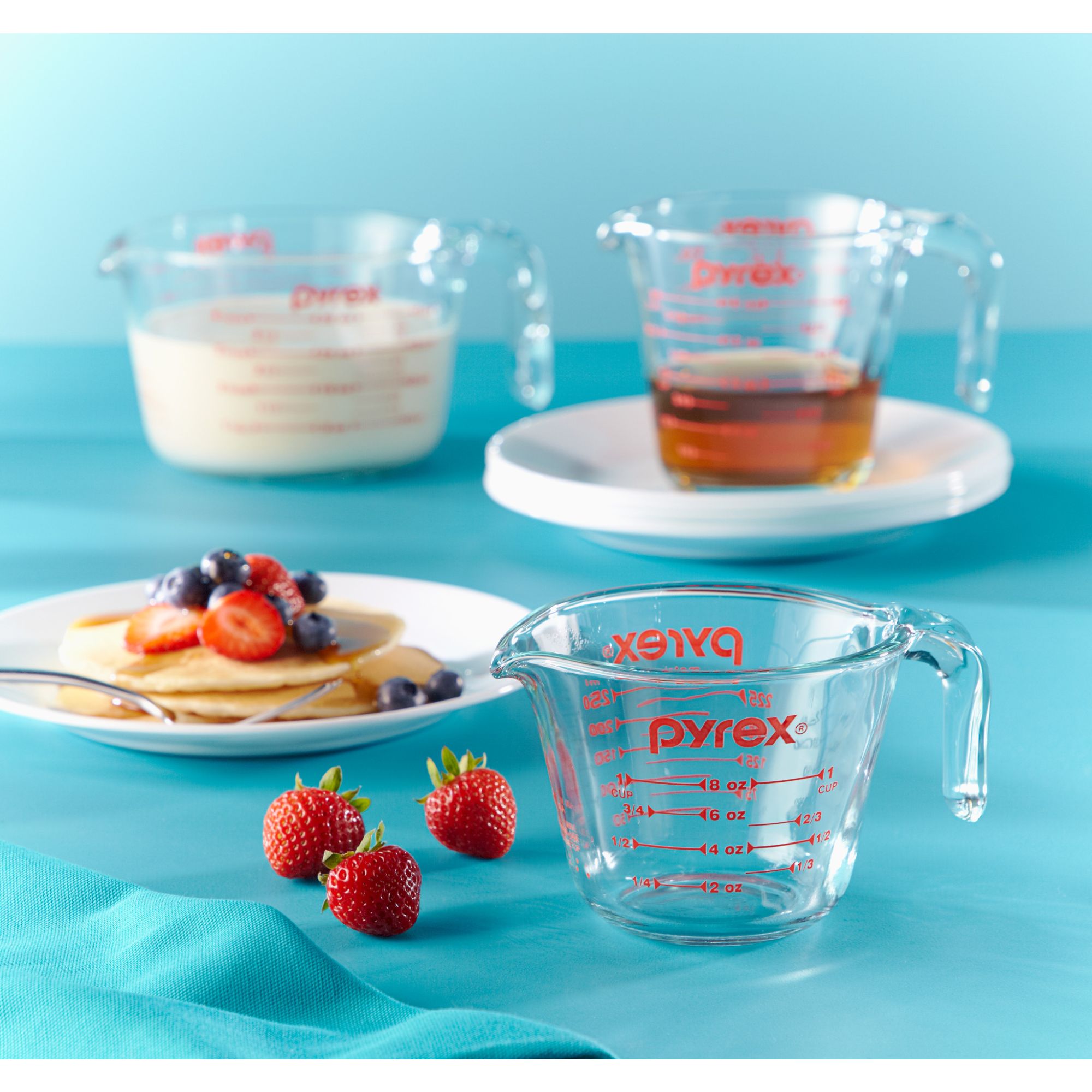 Photo of three Pyrex measuring cups. Background is blue. Image includes a plate with pancakes and berries on top with a fork. Strawberries on the blue table. Largest measuring cup contains milk, medium cup contains syrup, smallest cup is empty.