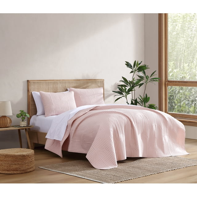 C&C California Embroidered Quilt Set, 100% cotton breathable shell with lightweight recycled polyester fill, 3-piece Full/Queen size in pink, featuring embroidered curved quilting patterns, displayed on a wooden bed with woven headboard in a bright bedroom with natural decor including a green plant, woven pouf, and neutral rug, ideal for year-round layering.