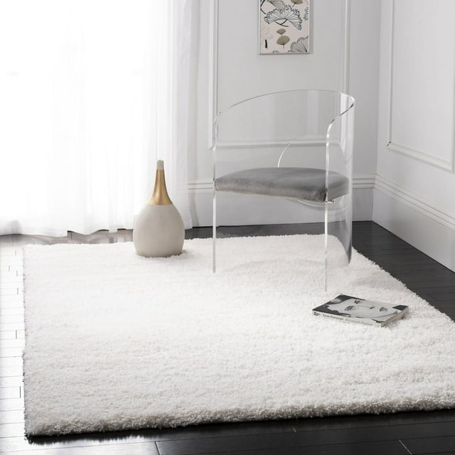 White Safavieh California Solid Plush Shag Area Rug displayed with a clear acrylic chair, modern vase, and book on dark wood flooring.