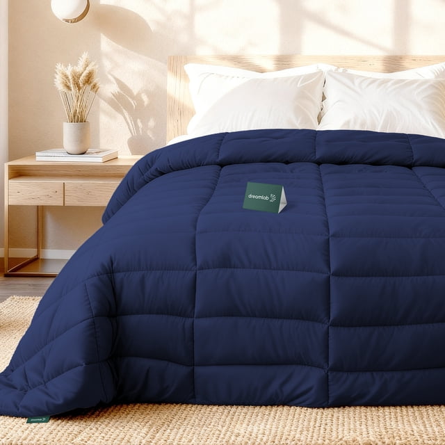 dreamlab edredón acolchado para toda estación. relleno de microfibras recubiertas con gel. lavado a máquina. usar como edredón o con funda duvet. hipoalergénico. (azul marino, queen 223 x 223cm) Edredón Queen azul marino, vista frontal centrada sobre cama con acolchado rectangular y costuras visibles que fijan el relleno; etiqueta Dreamlab y lazo de esquina visibles, superficie de microfibra de aspecto mate; fondo de dormitorio minimalista con mesita y almohadas blancas. También suele usarse sin funda como colcha ligera.