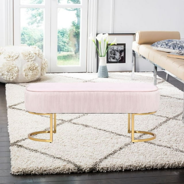 Oval pink velvet backless bench with pleated upholstery and gold metal legs on a white patterned rug in a bright living room.