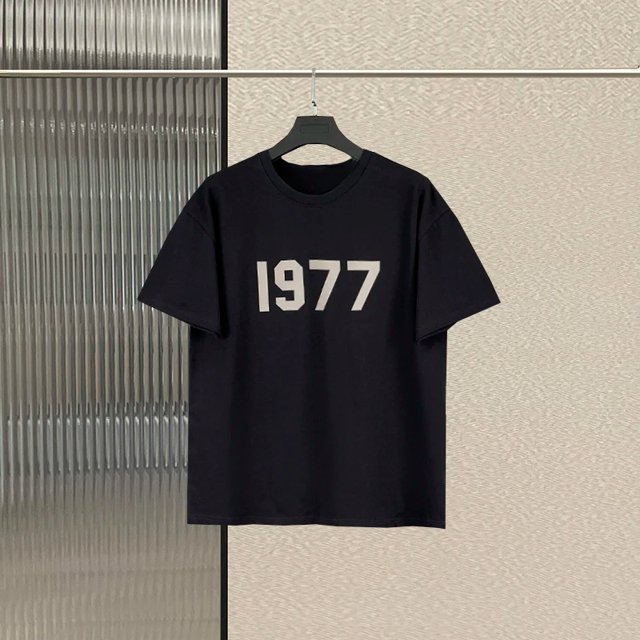 essentials-camisetas de manga corta con logo de letras para hombre y mujer, camisetas unisex de hip-hop, 1977 algodón, 100% Camiseta unisex negra de algodón con '1977' impreso en blanco en el pecho, vista frontal centrada colgada en percha, cuello redondo y mangas cortas, fondo interior minimalista; a menudo usada como prenda casual de estilo urbano.