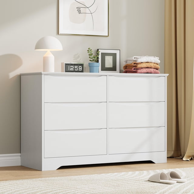 qinaqin cómoda de madera con 6 cajones, mueble alto moderno con cajones profundos y cubierta amplia, color blanco; vista frontal centrada de cómoda blanca de seis cajones con tiradores ocultos integrados en los frentes, base elevada con rebaje inferior y frentes de madera pintada que muestran líneas de separación que indican deslizamiento suave; superficie amplia con lámpara, reloj digital y ropa doblada, ambientación de recámara con cuadro y cortina. Adecuada para guardar ropa o juguetes y usar en recámara, sala o pasillo.