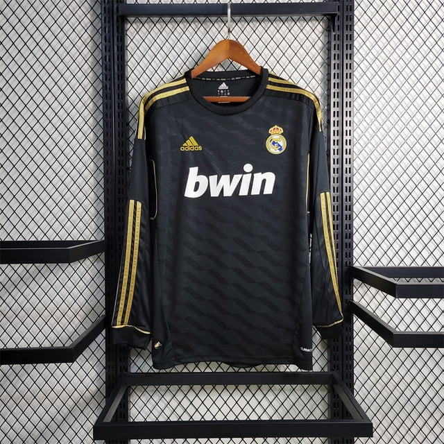 11/12 real madrid local/camiseta de fútbol visitante blanco/negro camiseta Real Madrid visitante negra con detalles dorados; vista frontal centrada colgada en percha, manga larga con tres bandas doradas de adidas en hombros y laterales, escudo del club en el pecho izquierdo y logo bwin blanco en el centro, tejido con patrón diagonal sutil. También suele usarse como camiseta de entrenamiento informal.
