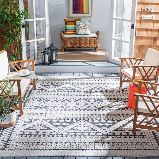 SAFAVIEH Cabana Arielle Geometric Indoor/Outdoor Area Rug in black and white with diamond and floral motifs, displayed on a sunlit porch with wooden frame chairs with white cushions, a red ceramic side table, potted plants, and open French doors leading to a wicker bench with colorful pillows. This durable outdoor rug is ideal for busy indoor or outdoor areas like decks and patios.