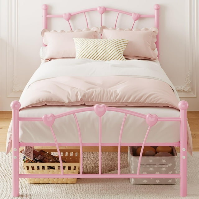 Metal Platform Twin Bed Frame Mattress Foundation with Iron-Art Sweat Heart Headboard & Footboard in pink. The bed frame features a heart-shaped design on the headboard and footboard, crafted from powder-coated metal for durability. It measures 78.7 inches in length, 39.3 inches in width, and 42.5 inches in height, with a 12-inch clearance underneath for storage. The frame supports up to 600 pounds and includes reinforced steel slats and a seven-leg construction for stability. Shown in a bedroom setting with soft pink bedding and storage baskets underneath.