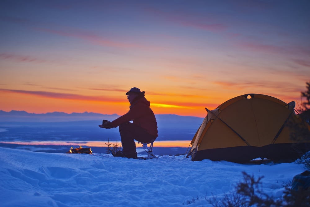 A backpacker warms his hands over a campfire at a campsite high in the Chugach Mountains overlooking Kinik Arm and Cook Inlet with Sleeping Lady in the background Southcentral Alaska Canvas Art - Mich