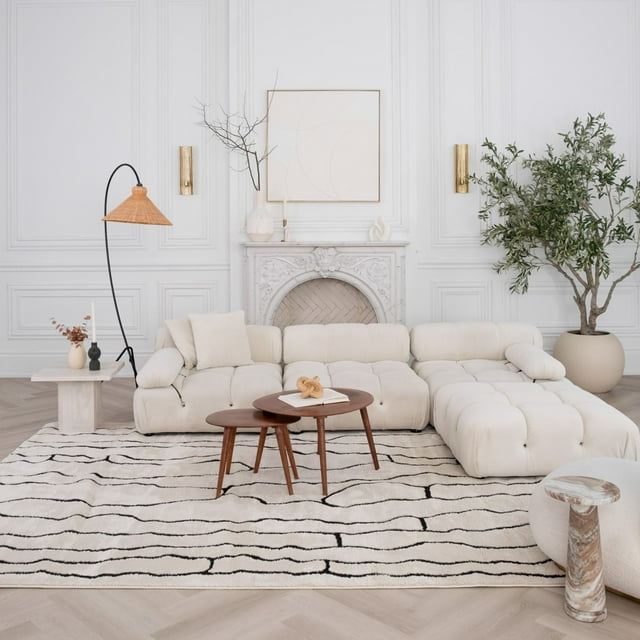 Modern cream and black abstract indoor area rug placed under a beige tufted sectional sofa with two wooden coffee tables, set in an elegant living room featuring white paneled walls, decorative fireplace, floor lamp, and large potted plant.