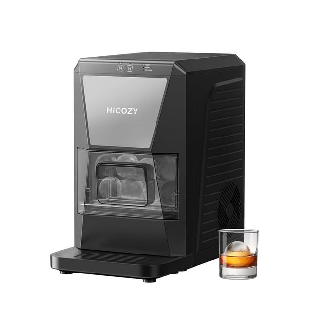 HiCOZY Ice Maker Clear Ice Ball Maker Countertop with IcyGem Technology, shown in a sleek black design. The machine produces two crystal-clear ice balls in 60 minutes and up to 40 pieces per day. The front-facing view highlights the transparent ice compartment containing round ice balls. A glass of whiskey with a single ice ball is placed beside the machine, emphasizing its use for cocktails, whiskey, and bourbon. Includes self-cleaning functionality and stainless steel ice tongs for convenience.