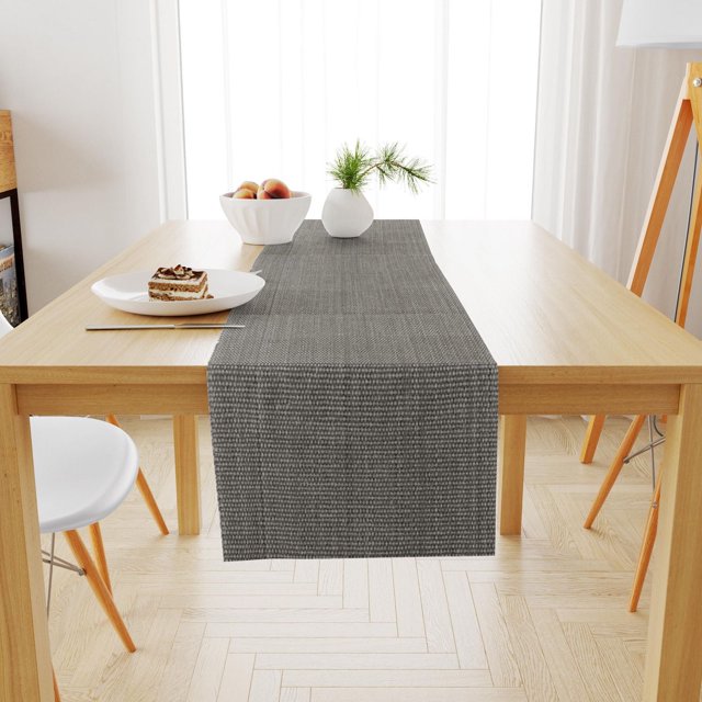 Gray woven cotton table runner on light wood dining table set in bright room with white bowl of peaches, small vase with greenery, and plate holding dessert.