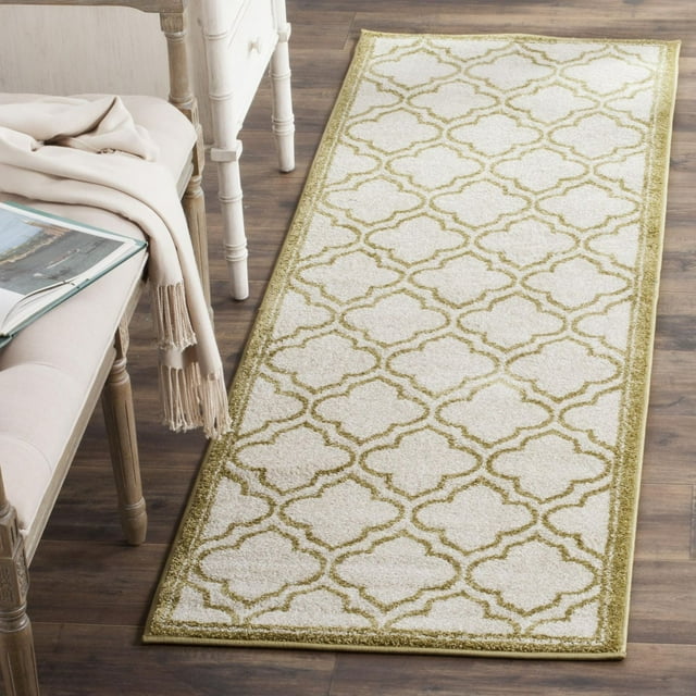 Safavieh Amherst Wilson geometric area rug in beige with olive green pattern displayed on wooden floor beside a chair draped with a light brown throw.