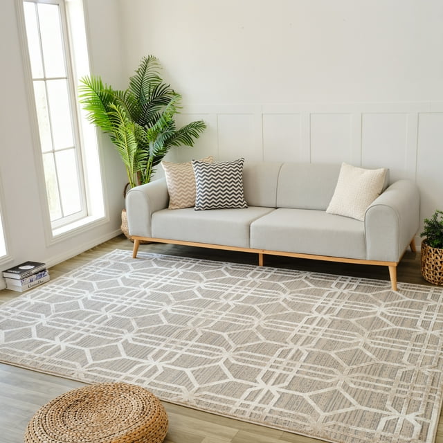 Modern beige and brown abstract indoor area rug with geometric pattern placed under a light gray sofa featuring chevron and textured pillows, complemented by green plants in woven baskets near large window.
