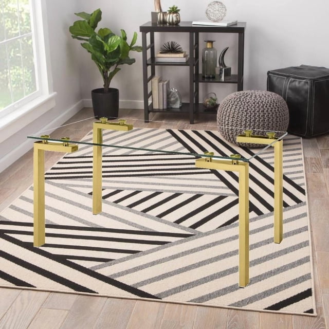 Modern rectangular dining table featuring gold stainless steel legs and a clear tempered glass top, placed on a black, white, and gray geometric rug in a bright room with wooden flooring.