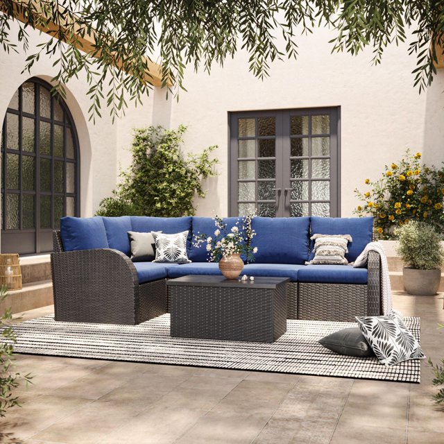CorLiving Brisbane 6-piece rust-resistant outdoor wicker patio sectional set with blue cushions and matching coffee table on a striped outdoor rug in a garden setting.