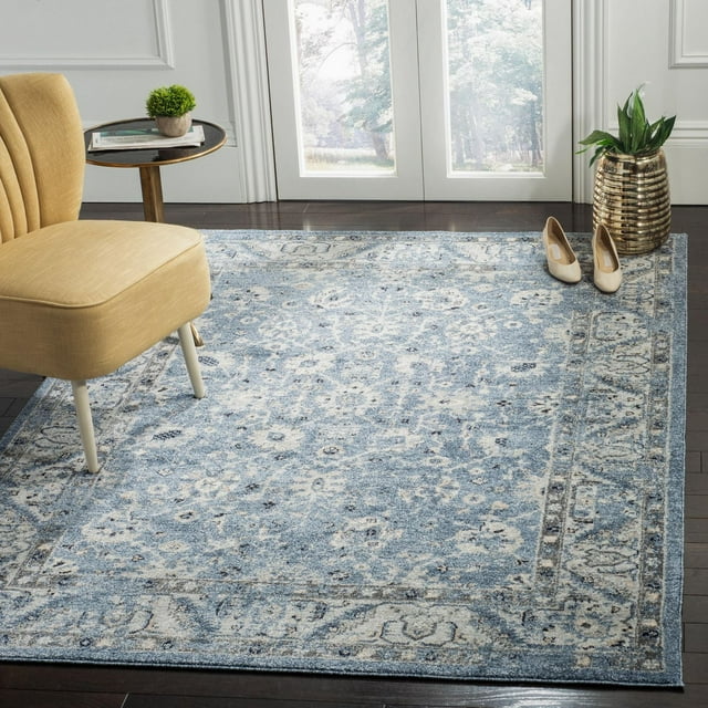 SAFAVIEH Charleston Tangier Overdyed Floral Area Rug in blue and cream tones displayed in a room setting with dark wood flooring, yellow upholstered chair, small round side table with books and plant, beige shoes near gold planter