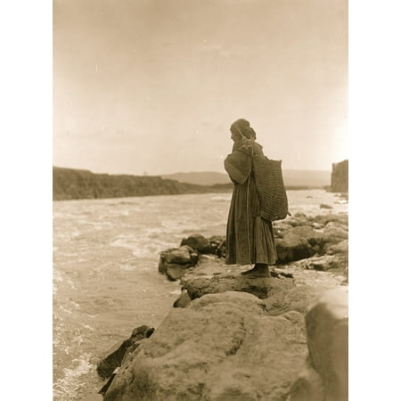 Tlakuit Indian standing on rock at waters edge wearing ankle length dress and large bag on her back strap supported on crown of her head Poster